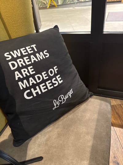The image shows a black cushion with white lettering "SWEET DREAMS ARE MADE OF CHEESE" and "Le Burger" placed on a green cushion and a beige seat. Part of a window can be seen in the background. The image was not taken in Amstetten, Austria.