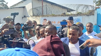 The image depicts a crowded outdoor scene in Kabinzi, Rwanda, where a group of approximately twenty to thirty adult males is gathered, seemingly for a press event or public address. In the immediate foreground, a man with dark skin and short hair is partially obscured by a maroon-colored knitted garment draped over his left arm, his gaze directed downwards. To his right, another man in a blue blazer over a white t-shirt looks forward, with a white handheld microphone positioned towards him. Surrounding these central figures, numerous individuals are actively documenting the event. Several men hold smartphones, some mounted on gimbals or selfie sticks, while others operate professional video cameras. One man wears a dark cap with a visible United Nations (UN) logo. In the background, a white banner is displayed against a wall, featuring text in French including "16 Jours d'activisme contre les violences faites aux femmes" (16 Days of activism against violence against women) and a graphic of hands covering a face with a "STOP" symbol, along with a UN emblem. Behind the banner, a light-colored building with a corrugated metal roof is visible, separated by a wall topped with multiple coils of concertina wire. The scene occurs during daylight hours.