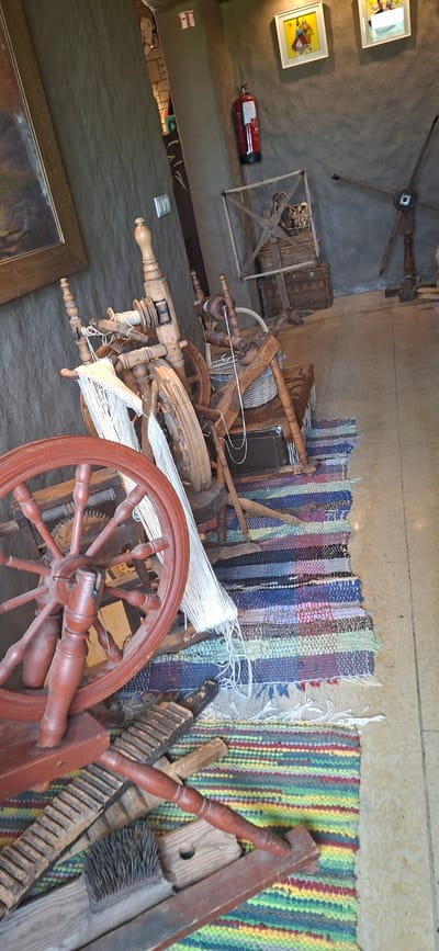 The image shows an interior scene of what appears to be a rustic exhibition or workshop, likely in Karja, Estonia. In the foreground, a large red-brown spinning wheel with a prominent rim is positioned to the left. To the right and slightly behind it, another wooden spinning wheel with a basket is visible. Between these, a colorful woven rug with stripes of various colors and frayed edges lies on the floor. A variety of antique tools, including what seem to be washboards and a brush, are scattered on the rug in front of the spinning wheels.

In the background, more wooden artifacts, possibly related to textile production, are present. A red fire extinguisher is mounted on a rough, textured wall to the upper left. Two framed artworks hang on the wall above the fire extinguisher. The walls have a rough, textured finish, suggesting an older or traditional building style. The lighting is somewhat dim, creating a slightly shadowy atmosphere. The overall impression is of a display of historical craft equipment in a rural or heritage setting.