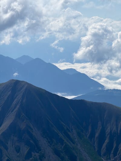 The image depicts a mountain landscape under a cloudy sky. In the foreground, a dark, steep mountain slope dominates the lower half of the frame, showing signs of erosion with visible gullies and patches of green vegetation. Behind this, several layers of blue-hued mountains recede into the distance. The sky is filled with a mix of bright white and darker gray clouds, with patches of blue visible. The scene conveys a sense of natural grandeur and depth. The location is Wildermieming, Austria. There are no people or specific objects visible in the image, only the natural environment.