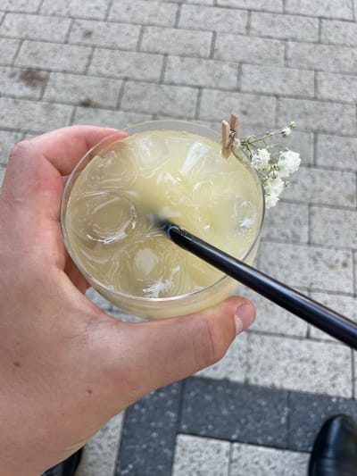 In Ratuș, Moldova, a person's hand holds a clear glass filled with a pale yellow, ice-filled beverage. A black straw protrudes from the drink, and a small, wooden clothespin secures a sprig of tiny white flowers to the rim of the glass. The scene is set against a backdrop of grey brick paving stones, indicative of an outdoor urban setting.