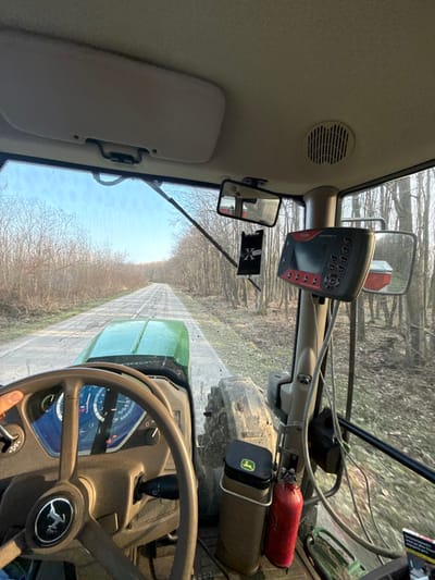 The image depicts the interior of a John Deere tractor from the driver's perspective, operating on a paved road in Slavianovo, Bulgaria. A gloved hand is visible on the brown steering wheel, which features the John Deere logo. The dashboard gauges, partially visible, indicate operation. Mounted on the right-side pillar is a precision agriculture display unit, possibly a Trimble GFX-750, with a red frame, grey body, and multiple control buttons. A rearview mirror and a "X" branded air freshener hang from the ceiling. Below the display, a brown cylindrical bin with a John Deere logo and a red fire extinguisher are secured. Through the windshield, a two-lane paved road, showing signs of wear, extends into the distance, bordered on both sides by dense, bare deciduous trees. The sky is clear blue, indicating fair weather. The right side mirror reflects a red structure, possibly an attached implement or trailer.