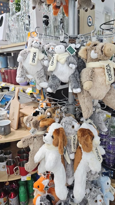 The image displays a retail setting, likely a gift shop or souvenir store, filled with a variety of stuffed animals. The stuffed animals are primarily on display racks, some hanging from hooks. The animals include what appear to be bears, dogs, and other fuzzy creatures, many wearing scarves. Several of the scarves have the word "CORSICA" written on them in black text, indicating a potential theme or origin related to Corsica, France. In the background, there are shelves stocked with various items, including what seem to be decorative bottles and mugs. The lighting suggests an indoor environment. There are no people visible in the image. The overall impression is a store showcasing merchandise, with a focus on Corsica-themed plush toys.