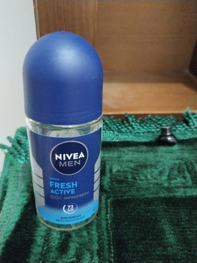 A close-up, slightly angled shot shows a Nivea Men Fresh Active roll-on deodorant bottle. The bottle is made of clear glass with a blue plastic cap and a blue label. The label features the Nivea Men logo in white, followed by "FRESH ACTIVE" in large blue text. Below that, it reads "∞ INFINIFRESH" and a circle with "72 JAM" inside. The bottom of the label has the text "Anti-bakteri ANTI-PERSPIRANT". The bottle is positioned on a dark green, plush, textured surface that appears to be a rug or fabric. In the background, there is a brown wooden surface and a light-colored wall. There is no indication of the city of Lahat, Indonesia in the image.