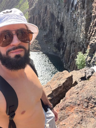 A man with a beard and sunglasses, wearing a white bucket hat and a backpack, is taking a selfie at the edge of a rocky cliff overlooking a body of dark water with sunlight sparkling on its surface. The cliff face is steep and rugged, with visible vegetation and a small waterfall on the right. The man is shirtless, wearing light-colored shorts. The overall impression is a scenic outdoor location, possibly a natural gorge or canyon.