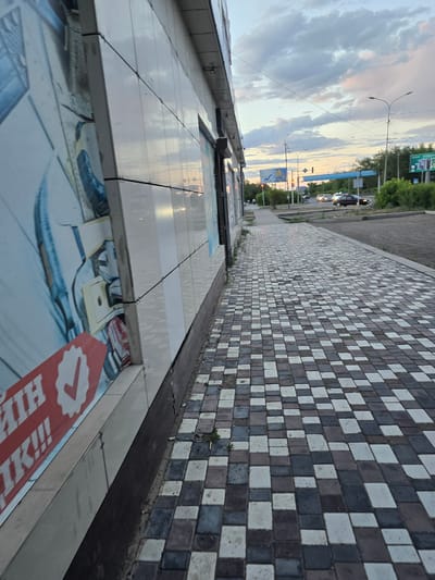 This is a street-level, eye-level, medium shot of a sidewalk in Satbayev, Kazakhstan, captured during the day under a cloudy sky. The photograph is taken from a low angle, looking down the length of a paved sidewalk. The sidewalk is constructed of interlocking square and rectangular pavers in a checkered pattern of black, white, and a reddish-brown color. To the left, a portion of a building's facade is visible, clad in light-colored tiles and adorned with a large graphic advertisement. The advertisement features a red banner with white text and a white checkmark logo. Beyond the sidewalk, a road is visible with a few cars driving. Trees line the far side of the road, and in the distance, a traffic light is visible, indicating an intersection. The overall scene depicts a typical urban environment.