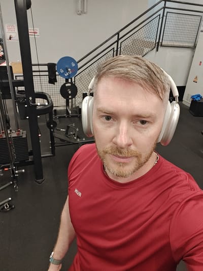 An adult male is depicted in a close-up selfie taken within a gym facility in Nahariya, Israel. The man has fair skin, light brown hair swept to the right, and a reddish-blonde beard and mustache. He is wearing a red short-sleeved athletic shirt with "ЯUR" (likely "RUN" in reverse) printed in white on the left chest, a silver-colored chain around his neck, and white over-ear headphones. A light blue/green patterned watch strap is visible on his left wrist. He is looking directly at the camera.

The background reveals a gym environment with a dark, possibly rubberized floor and light-colored walls. On the left, a black cable machine with visible weight stacks is present. In the mid-ground, a barbell rack or similar equipment features a blue weight plate marked "50" and a black weight plate. A black metal staircase with a grid-patterned railing ascends in the right background. A white sign on the left wall contains red Hebrew text, including "לתרגיל הבא" (To the next exercise) and "הרמת רגל" (Leg raise), accompanied by a graphical representation of an exercise. Overhead lighting fixtures are visible. A blue cloth object rests on a surface in the far right corner.