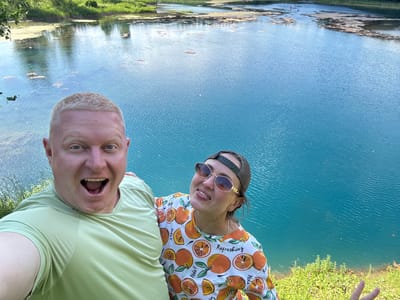 This is a selfie of two people, a woman and a man, posing in front of a body of bright blue water, possibly a lake or a river. The image appears to be taken outdoors on a sunny day, as indicated by the bright lighting and the reflections on the water. The woman is on the left, wearing a light-colored shirt with an orange and white fruit pattern and sunglasses. She is smiling and sticking out her tongue. The man is on the right, wearing a light green polo shirt and has his mouth open in a surprised expression. Both individuals are positioned upside down relative to the water and the background, suggesting they have either rotated the camera or themselves for the photo. The background shows lush green grass and vegetation along the edge of the water, and the water itself is calm with visible ripples. The reflection in the water shows the sky and hints of trees and a grassy shore. The scene is set in a natural environment. The shirt the woman is wearing has text that reads "Refreshing" and "Hola!!".