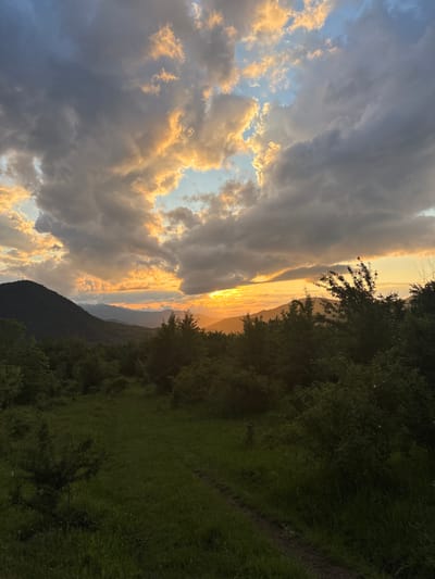The media file captures a dramatic natural landscape at either sunrise or sunset in Iamna, Bulgaria. The sky dominates the upper portion of the scene, filled with a dynamic blend of clouds. Large, dark grey clouds occupy much of the frame, contrasting sharply with patches of bright, golden-orange illuminated clouds where the sun's rays break through. Glimpses of clear blue sky are visible in between the cloud formations.
On the right side and across the lower part of the image, the terrain consists of dense, dark green trees and lush grass. These elements appear largely as silhouettes due to the strong backlighting from the low sun. Individual tree branches with discernible leaves extend into the illuminated sky. In the mid-ground, a range of mountains or hills stretches across the horizon, forming dark, undulating silhouettes against the intense, warm glow of the setting or rising sun. The sun itself is partially visible along the distant horizon, casting a fiery, golden light that defines the landforms and highlights the cloud edges.
No people, animals, or specific man-made objects are visible. The image focuses entirely on the natural environment and a spectacular display of atmospheric light. The time of day is clearly indicated as either dawn or dusk by the warm, intense illumination and the play of light and shadow. The weather appears to be partly cloudy with significant cloud cover, but with breaks allowing for this dramatic light show. The mountainous terrain and dense greenery are consistent with the provided location context of Iamna, Bulgaria. There is no visible text in the image. The overall impression is one of serene natural grandeur and powerful beauty.