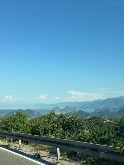 The image presents a view from a roadside location in Gornja Brova, Montenegro. The foreground shows a gray metal guardrail along a road. Beyond the guardrail, a landscape of mountains and water is visible. The sky is clear and blue. The mountains in the distance are hazy and pale in color. The roadside vegetation includes green trees and grass. The overall perspective is looking down from a high vantage point.
