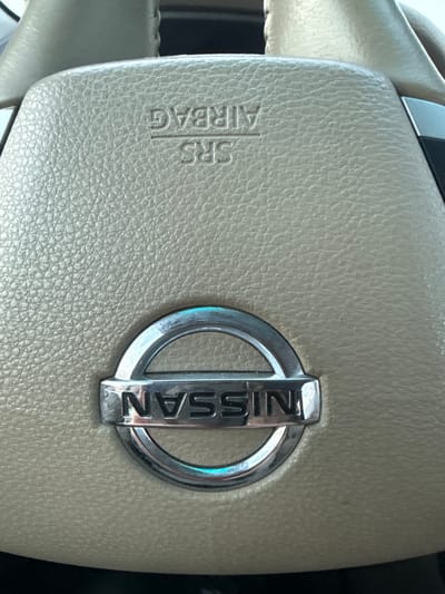 The image displays a close-up, head-on view of the central section of a vehicle's steering wheel, specifically within a car located in Bilimbay, Russia. The steering wheel material is a light beige or tan, textured surface, likely synthetic leather or plastic. Centered on the steering wheel is a chrome Nissan logo, featuring the word "NISSAN" horizontally within a rectangular bar, which is itself enclosed by a circular chrome ring. Minor scuff marks and reflections are visible on the chrome logo. Above the Nissan emblem, embossed into the steering wheel's surface, are the words "SRS" and "AIRBAG", both oriented upside down relative to the Nissan logo. "SRS" appears above a subtle horizontal line, and "AIRBAG" appears below it. No individuals, specific actions, or further environmental details beyond the immediate vehicle interior are discernible.