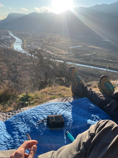 A high-angle shot reveals people sitting on a blue quilt atop a hill, overlooking a valley with a river. Hiking shoes and casual pants are visible. A small black JBL speaker sits on the quilt, along with a blue object. A hand holds a rolled cigarette. The background shows a mountainous landscape under a bright, sunny sky. Bare trees and vegetation dot the hillside.