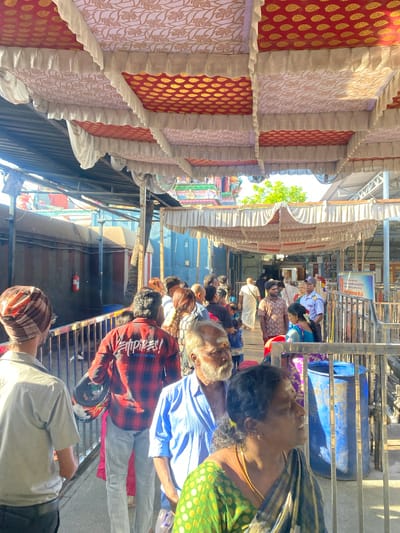 The image captures a view from within a covered walkway at a temple in Chinnambedu, India. Overhead, decorative canopies with red and gold patterns, edged with cream fabric ruffles, are visible. In the foreground, a group of people are standing in a line, with metal railings separating them from an area to their right.

A man in a light-colored shirt and a patterned turban is on the far left, facing away from the camera. Next to him, a person in a red and black plaid shirt holds a helmet. A man with a white beard wearing a blue shirt is in the center foreground, looking to his right. To his right, a woman in a green and black sari is also looking to her right.

Behind the people, a long, dark structure runs along the left side of the walkway, with a red fire extinguisher mounted on it. The right side of the walkway features more railings and a large blue cylindrical container. In the background, glimpses of temple architecture and greenery can be seen. The scene appears to be an outdoor queuing area, likely for entry into a temple. The lighting suggests it is daytime, with sunlight filtering through the canopy.