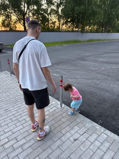 In Kraskovo, Russia, a man and a young child are in an outdoor parking lot. The man, wearing a white t-shirt, black shorts, and sneakers, is facing away from the camera. A black strap from a bag is visible over his shoulder. The child, dressed in a pink t-shirt and blue striped shorts, is bent over, looking at a silver bollard with red stripes. The scene is set on a paved area next to a grassy strip and trees. The lighting suggests it is late afternoon or early evening, with warm sunlight filtering through the trees. The mood appears relaxed and casual. No visible text is present in the image.