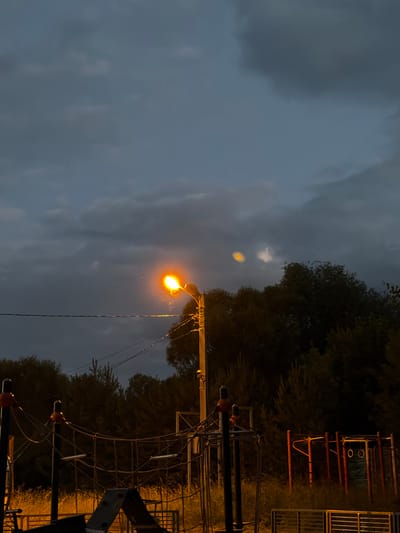 The image depicts a dimly lit outdoor scene at dusk or night, likely in the city of Martemyanovo, Russia. A single street lamp illuminates a portion of a playground or outdoor fitness area. The sky is overcast with dark clouds, and the light from the lamp casts a warm glow on its surroundings, highlighting the metal structures of the playground equipment, including ropes and bars. Trees form a dark silhouette in the background. The overall atmosphere is subdued and quiet.