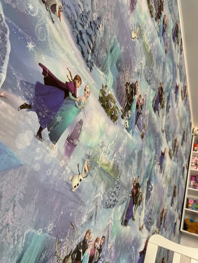 The image shows a wall covered in a "Frozen" themed wallpaper, featuring characters like Elsa, Anna, Olaf, Kristoff, and Sven in a snowy, icy landscape. The scene depicts the characters in various poses, some appearing to be ice skating, others riding on Sven. In the background, there are depictions of ice castles and snowy trees. On the right side of the image, a portion of a white bookshelf is visible, containing various items, some of which appear to be children's toys or craft supplies. The lighting suggests an indoor setting. The image is a close-up view of the wallpaper, with the angle emphasizing the repeating pattern. There is no discernible location in the city of Мариинский, Russia, as this is an interior shot of a decorated wall.