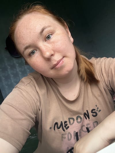 A close-up, head-and-shoulders selfie shows a young Caucasian woman with light skin, freckles, and light red hair tied in a ponytail. She has light blue eyes and is looking directly at the camera. She is wearing a light brown t-shirt with black lettering that reads "MEDONS FORCED". The background is dark and appears to be indoors, with a textured wall visible on the left. There is no indication of the city of Аряж, Russia within the image.