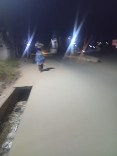 At night, a woman walks along a sidewalk with a load balanced on her head, moving away from the camera. Streetlights shine brightly nearby. The sidewalk is next to a dirty drainage ditch filled with debris. The street is wide and empty, leading into the dark background. The overall scene is dim with poor visibility.