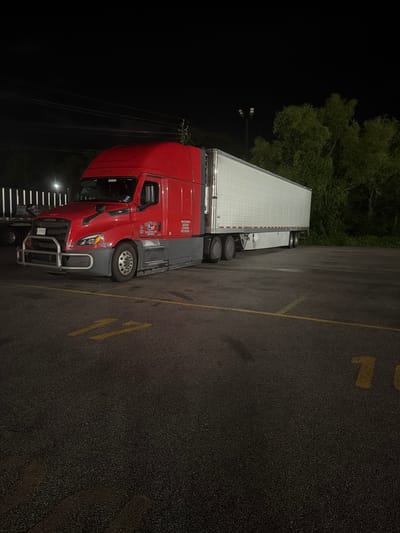 A red and grey semi-truck is parked in an asphalt lot at night. The truck is positioned at an angle, facing towards the left side of the frame. The trailer is white and has a long, rectangular shape. Yellow painted numbers are visible on the asphalt in the foreground, indicating parking spaces. The background features trees and utility poles with lights. The sky is dark. There is no visible activity or interaction in the scene. The location is identified as Wildwood, United States.