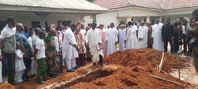 People gather beside a grave. Some wear traditional clothing, others are in white religious garb. One man in white stands near the edge of the grave with a book in hand. Another man uses a shovel to put dirt into the grave. The surrounding area appears damp, and there is a building with white columns in the background. The scene appears to be an outdoor funeral ceremony.