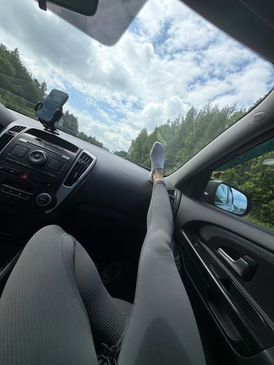 The image, oriented diagonally, captures the interior of a car from the perspective of a passenger. The lower body of a person is prominent, wearing light gray, ribbed leggings and white socks, with one foot casually propped on the dark dashboard.
The car's interior features a dark dashboard, a central console with a radio/media system (buttons like "FM," "AM," "CD," "AUX," "SCAN," "A/C" are visible), air vents, and a hazard light button. A smartphone in a black car mount is affixed to the dashboard, displaying "95" on its screen, which could indicate speed or a road number. A bottle is partially visible in the door's storage compartment. The passenger-side A-pillar has an "SAS Airbag" label.
Beyond the car's confines, a bright, cloudy blue sky dominates the view through the windshield. Lush green trees line what appears to be a road or forest, and distant structures or poles are faintly visible. A yellow road sign is also barely discernible in the distance. The outdoor scenery reflected in the side mirror further confirms the forested environment. The bright natural light suggests it is daytime. The overall scene depicts a relaxed moment during a car journey, likely through a rural or wooded area near Башкултаево, Russia.