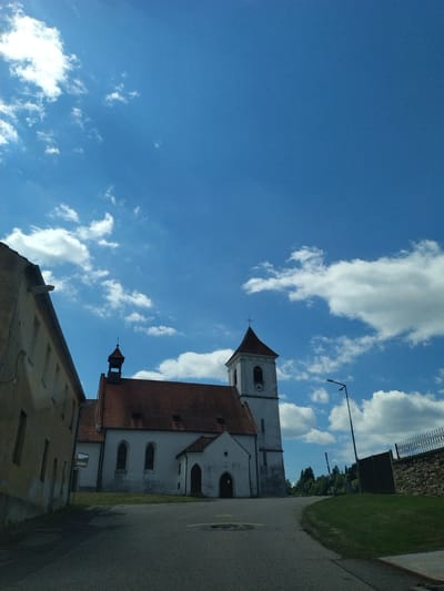 The image shows a street scene in Květušín, Czechia, featuring a church and adjacent buildings. The sky is a vibrant blue with scattered white clouds. On the left, a light-colored building with a sloping roof and several windows is visible. To the right of this building is a white church with a red tiled roof. The church has a tall, square bell tower with a pointed roof, a clock face, and a cross at its apex. A smaller entrance structure with a dark arched doorway is attached to the main church building. The street in the foreground is paved and appears to be an asphalt road. To the right of the road, there is a grassy embankment leading up to a stone wall topped with a black metal fence. Trees and a lamppost are also present in the scene. The overall impression is a sunny day in a quiet European village.