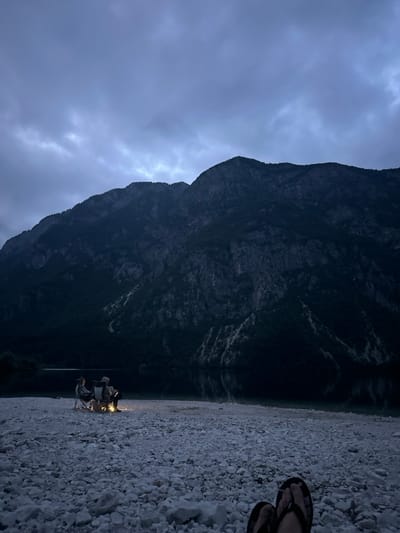 An overcast sky contrasts with a dark mountain, its base meeting a still body of water reflecting the mountain's silhouette. On a pebble-strewn shore, a small group of people sit around a campfire, tiny lights against the encroaching darkness. The feet of a person wearing sandals are visible in the foreground, adding a sense of scale and personal presence to the vast landscape. The scene conveys a feeling of quiet solitude and the intimacy of shared moments.
