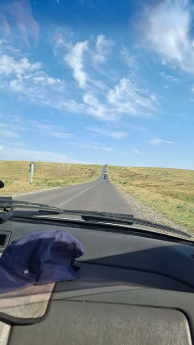 The image is a POV shot from inside a vehicle, looking forward. A blue baseball cap rests on the dashboard in the foreground, casting a shadow. The vehicle is on a paved road that curves towards a distant, rolling hill under a bright blue sky with scattered white clouds. The landscape on either side of the road appears to be dry, grassy fields. Several other vehicles are visible in a line receding up the road, suggesting a convoy or slow traffic. Utility poles are intermittently spaced along the side of the road and on the hill. A road sign is present on the left side of the road near the foreground. The reflection of a hand wearing a ring is visible in the upper portion of the windshield. The scene is likely in a rural or semi-rural area, given the open landscape and the road infrastructure. The location is identified as Boraldai, Kazakhstan.