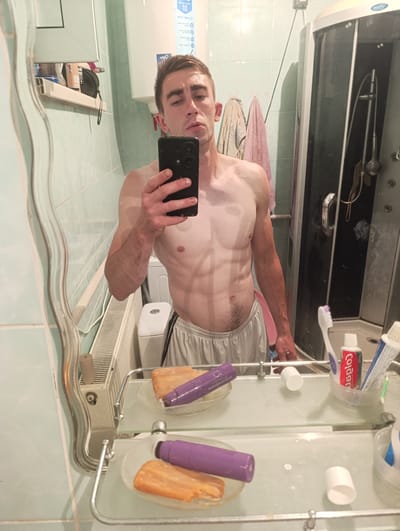 The media file depicts a man taking a mirror selfie in a bathroom. The man appears to be in his early to mid-twenties, with short, dark hair and a lean, muscular physique, visible through his bare chest and toned abdomen. He is wearing light grey athletic shorts with black trim. He is holding a black smartphone in his right hand, using it to capture the reflection.
The bathroom setting is somewhat utilitarian, with pale green tiled walls. To the left, a large mirror with a slightly distorted reflection is prominent, reflecting the man and part of the room. Behind the man, a white electric water heater is visible. To his right, a glass-enclosed shower stall is present, with a showerhead and handle visible. A washing machine is also partially visible in the background.
On the mirrored shelf below the man, several toiletries are arranged, including a bar of orange soap, a purple bottle or tube, toothpaste (Colgate is visible on the tube), and a toothbrush. A small white cap or lid is also on the shelf.
The lighting suggests it is likely daytime, but the indoor setting makes it difficult to determine the exact time of day or weather. The man's expression is neutral. The overall impression is of a casual, personal moment captured in a functional bathroom space. There is visible text on the water heater and the toothpaste tube.