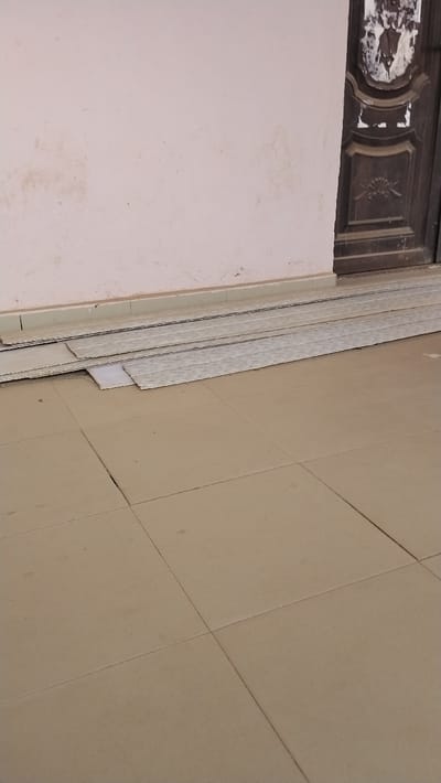 The scene depicts an exterior or semi-exterior building area in Ezzamgbo, Nigeria. The foreground features a light brown tiled floor with visible grout lines. Against a light pink wall, a stack of multiple rectangular, light-colored panels, possibly ceiling or wall cladding, rests horizontally on the tiled surface. The wall exhibits dirt marks and discoloration. To the right, a dark brown wooden door with decorative carvings is partially visible. The upper section of the door shows signs of wear, including torn or peeled material. No individuals or active events are present.