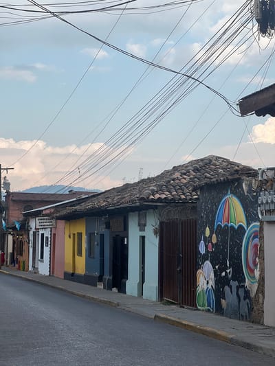 The image depicts a street scene in La Esperanza, Honduras. A row of colorful buildings lines the right side of the street, featuring hues of white, pink, yellow, blue, and light blue. The buildings have tiled roofs and a mural of umbrellas. Above, a dense network of electrical wires crisscrosses against a backdrop of a cloudy sky. A street runs along the side of the buildings.