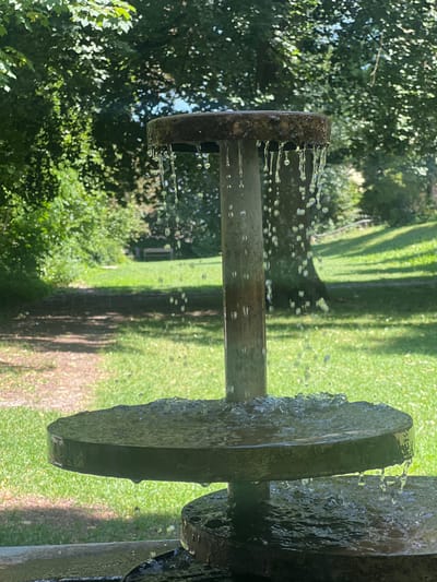 The image shows a close-up of a tiered fountain with water cascading down from the upper tier to the lower tier. The fountain appears to be made of metal and is positioned outdoors in a grassy park setting with trees in the background. The water is captured in mid-fall as droplets and splashes. The environment suggests a sunny day with dappled light filtering through the leaves. The fountain is likely located in the city of Moosburg, Germany, a place known for its parks and green spaces. There are no people visible in the image.