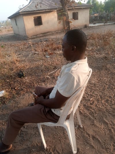 A man sits on a white plastic chair outdoors, holding a phone. He wears a short-sleeved shirt and brown pants, with dark sandals. The ground is sandy and sparsely vegetated. Behind him stands a single-story, aged house with a corrugated roof and small, shuttered windows. A few other buildings are partially visible in the distance. A dry tree stands close to the house. The scene is bathed in soft, warm sunlight.