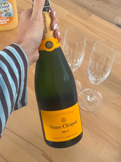 A person holds a bottle of Veuve Clicquot champagne, poised to pour. Two empty champagne flutes sit on a wooden surface. The bottle's yellow label is prominently displayed. A container of orange juice is partially visible in the corner. The person is wearing a striped shirt with brown and light blue stripes. The overall scene suggests a celebratory occasion or a brunch setting.