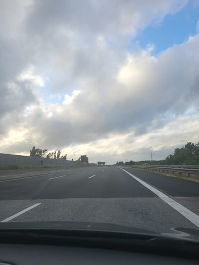 The image shows a view from the perspective of a vehicle driving on a multi-lane highway in Padenstedt, Germany. The sky is filled with a mix of dark and light clouds, with patches of blue sky visible. The highway is paved and marked with white lines. On the left side of the highway, there is a sound barrier and some trees. On the right side, there is a guardrail and a dense line of trees. There are a few other vehicles visible in the distance on the highway. The overall impression is of a cloudy day on a highway.