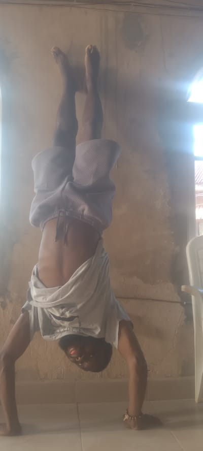 A person is performing a handstand indoors, presumably in Bende, Nigeria. The person has their feet positioned upward against a wall near a window, and the window is allowing natural light into the room. The person is wearing light-colored shorts and a t-shirt that is draped over their torso due to the inverted position. Their hands are on the tiled floor. A white plastic chair is visible in the background.
