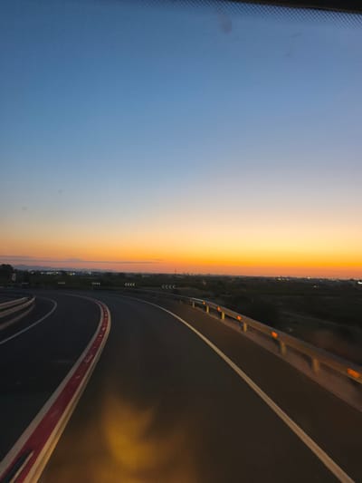 The image depicts a highway with a clear horizon at dusk. The sky transitions from a clear blue at the top to a vibrant orange and yellow near the horizon. In the distance, a city skyline with lights is visible. The highway itself is asphalt with white and red markings. Guardrails line the side of the road, and some have orange reflectors. The scene appears to be a view from a vehicle traveling on the highway. There are no people or animals visible in the image. The location is identified as the city of Alcàsser, Spain, likely on a highway leading into or out of the city. The time of day is clearly sunset or twilight.