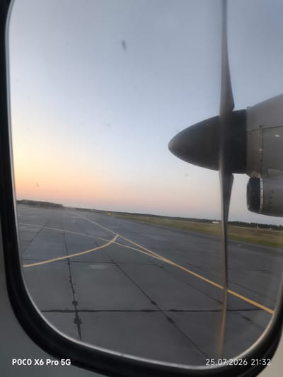 This image is taken from the inside of an airplane window, looking out at an airport runway at dusk. The propeller of the aircraft is partially visible in the foreground, obscuring some of the view. The runway is paved with asphalt and marked with yellow lines. In the distance, there is a treeline and the sky is a gradient of pale blue and orange, indicating sunset or sunrise. The date and time stamp on the image indicates it was taken on July 25, 2026, at 21:32. The text "POCO X6 Pro 5G" suggests the image was captured with that model of smartphone. There is no information in the image that allows for determination of the specific location within the city of Kogalym, Russia.