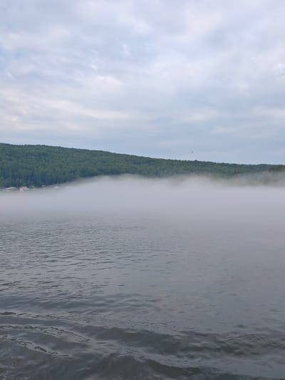 The image depicts a serene, misty morning scene over a body of water, likely the Yenisei River, in the vicinity of the village of Atamanovo, Krasnoyarsk Krai, Russia. In the foreground, the water surface is characterized by gentle ripples and a dark, grey-blue hue. The middle ground is dominated by a low-lying bank covered in dense green forest, which gradually slopes upwards. A thick layer of white fog hovers just above the water's surface, obscuring the base of the forested bank and lending an ethereal quality to the scene. On the left side of the middle ground, several small houses with visible roofs are partially visible through the mist, suggesting a settlement along the river. The sky above is overcast with a pale grey, textured cloud cover, with hints of a lighter blue peeking through in some areas. A small dark silhouette, possibly a bird in flight, is visible against the sky in the upper right quadrant. The overall atmosphere is tranquil and cool, indicative of early morning fog.