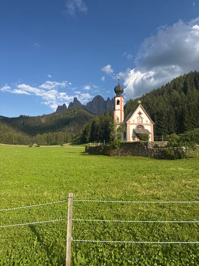 This is a vertically oriented, eye-level shot of the St. Magdalena church in the Italian Alps. The church, with its characteristic red and white striped bell tower and ornate facade, is nestled against a backdrop of dense evergreen trees and dramatic, jagged mountain peaks. A bright blue sky with scattered white clouds dominates the upper left portion of the image. In the foreground, a vibrantly green, well-maintained grassy meadow stretches across the right side, separated from the church and trees by a low, rustic stone wall. Several strands of white, twisted rope, supported by a wooden post, run horizontally across the meadow, suggesting a boundary or perhaps part of a temporary fence. The sunlight suggests it is daytime, likely mid-morning or afternoon, casting subtle shadows and illuminating the landscape. The overall impression is one of serene beauty and picturesque alpine scenery. No people or text are visible.