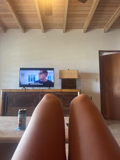 The image depicts an indoor scene within a residence in Olde Yard Village, British Virgin Islands, captured from a first-person perspective. In the immediate foreground, the bare, tanned legs of an individual are visible from the upper thighs to just above the knees, positioned slightly apart. Between the legs, on a light-colored, textured surface, sits an open aluminum can, primarily blue and white, with the visible text "Delight Iced Coffee Mocha."
In the midground, a dark-colored flat-screen television is centered on a long, dark wooden cabinet or credenza featuring bamboo-style trim. The television screen displays a man wearing glasses and a dark shirt, with white subtitles reading "Are you okay, ... wanting to be." The background on the television screen shows white architectural elements with blue window frames. To the right of the television, a table lamp with a rectangular, light-colored fabric shade and a dark, textured base rests on the cabinet. The wall behind these objects is a plain, light off-white color.
In the background, to the far right, a partially open brown wooden door reveals a glimpse of a light-colored wall in an adjacent room. A light switch or electrical outlet is visible on the wall to the right of the lamp. The ceiling features exposed wooden beams and lighter wooden planking, with a dark-colored ceiling fan, incorporating a light fixture that emits a warm glow, visible towards the top center. No direct human interaction is depicted; the scene suggests a person relaxing while viewing television.