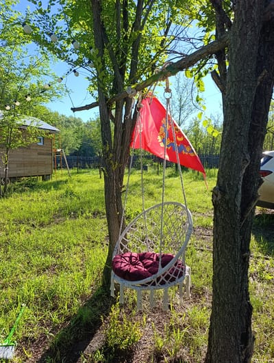 In Dimitrovskii, Russia, a white macrame hanging chair with a burgundy cushion is suspended from a tree branch. A red flag with a golden emblem is draped over the same branch, partially obscuring the chair. The scene is set outdoors in a grassy area with trees and a wooden building visible in the background. String lights are also strung across the trees.