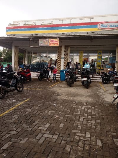 The image shows a storefront of a store called "Indomaret" in Kecamatan Caringin, Indonesia, located at latitude -7.523 and longitude 107.485. There are six motorcycles parked in front of the store, and several people are standing around, some of them are wearing helmets. There is a blue trash can in the foreground, next to a yellow sign on the ground. The pavement is made of brick and is mostly covered in dirt. There is a sign above the storefront with the word "Indomaret" on it. The store has a large glass window, and there are shelves visible inside.