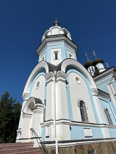 A low-angle, full-shot photograph captures the exterior of an Orthodox church against a clear, vibrant blue sky. The church is painted in a combination of white and light blue, with black accents on the roofs and window frames. A prominent dome, topped with a golden cross, rises towards the sky. The church features multiple windows with arched tops and decorative iron grilles. The building's architecture includes several tiered sections, decorative moldings, and a stone-clad foundation. To the left of the church, a tall, dark green conifer tree is visible. The foreground shows a set of reddish-brown steps leading up to the church entrance. The scene appears to be set in St. Ozerki, Russia, indicated by the architectural style and the presence of religious iconography.