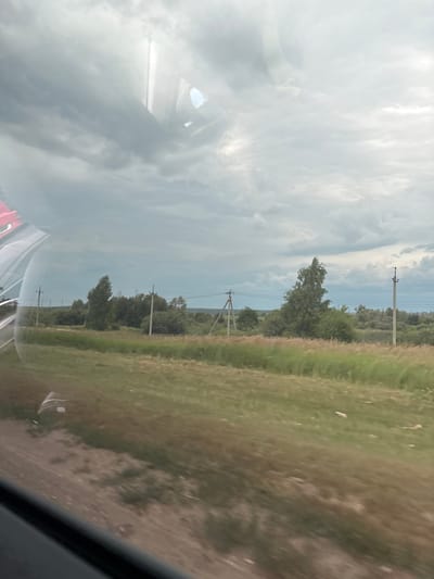 The image depicts a rural landscape seen through a window, likely from a moving vehicle. The foreground shows a dirt and grassy roadside. Beyond this, a field of tall grass stretches towards a treeline. Several utility poles with power lines are visible, interspersed with trees and bushes. The sky is overcast with dramatic, dark clouds, suggesting a stormy or approaching storm. The location is identified as Krasnaya Gorka, Russia. Reflections are visible on the window, including what appears to be part of the vehicle's interior and a side mirror.