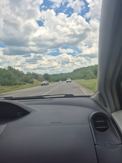 The image captures a view from inside a car traveling on a highway near Vadul lui Vodă, Moldova. The sky is filled with dramatic, billowing white clouds against a backdrop of blue sky. The highway stretches ahead, flanked by lush green trees and rolling hills, indicating a rural or semi-rural setting. Several cars are visible on the road, including a dark-colored sedan in the foreground and a white SUV further ahead. The interior of the car is also visible, showing the dashboard with a digital display indicating the time as 12:40 and a speed of 64 km/h. The overall scene suggests a pleasant, overcast day on a road trip through the Moldovan countryside.