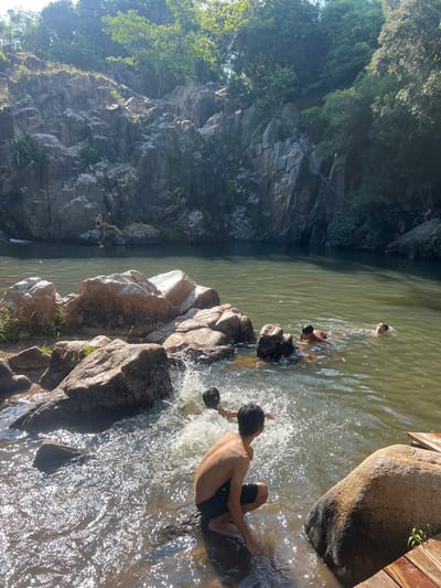 The image depicts a natural swimming area amidst a rocky landscape with lush vegetation. In the foreground, a young person is partially submerged in the water, appearing to crouch or sit near the edge. To their right, a large, smooth boulder sits on wooden planks. The water is disturbed by splashes, indicating movement and activity. In the middle ground, several other individuals are swimming or playing in the water. Further back, a rocky cliff face rises, with two people seated on rocks near its base. The water is a murky green, reflecting the surrounding trees and rocky formations. The scene is bathed in sunlight, with rays of light filtering through the trees, creating a bright and airy atmosphere. The location is identified as Vĩnh Hải, Vietnam.
