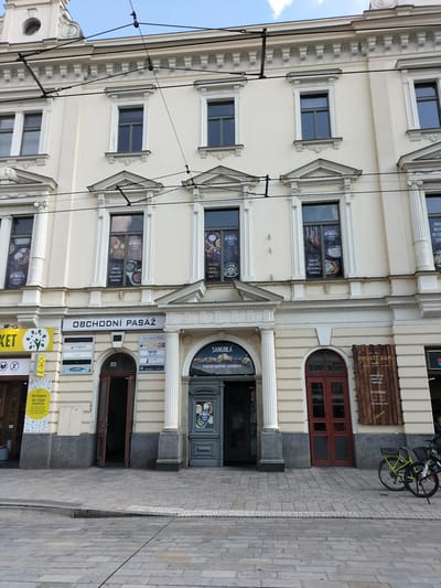 This is a photograph of the exterior of a building in Pardubice, Czechia, on a sunny day. The building has a light-colored facade with multiple windows and ornate architectural details. The ground floor features several shop entrances and signage.
Visible businesses and text include:
- "OBCHODNÍ PASÁŽ" (Shopping Arcade) above a set of entrances.
- Signs for businesses like "TOBRYS," "Kosmetika Stella," and "MAMA."
- A yellow sign with a green tree logo that says "MARKET."
- A yellow sign with text including "EKO drogerie BIO/VEGAN kosmetika."
- Signs for "SANGRILA," described as an "INDICKÁ NEPÁLSKÁ RESTAURACE" (Indian-Nepali Restaurant).
- A sign for "PROSTĚ BAR," advertising a cocktail bar, snack bar, and dance hall, with operating hours.
There are also decorative posters in some of the windows, advertising food and the restaurant. Two bicycles are parked on the sidewalk in front of the building. The scene appears to be a bustling street with pedestrian traffic, indicated by the paved sidewalk. The overall impression is that of a commercial street in a European city.