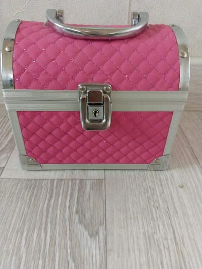 A pink, quilted vanity case with silver trim and a silver handle sits on a light-colored wooden floor. The case has a diamond-quilted pattern with subtle glitter embedded in the fabric. A silver latch with a keyhole is located on the front. The background is a textured white wall. The image is taken from a slightly elevated, straight-on angle. There are no people or discernible actions taking place. The scene appears to be indoors. The location of Prigorognoye, Kyrgyzstan is not visually indicated in the image.