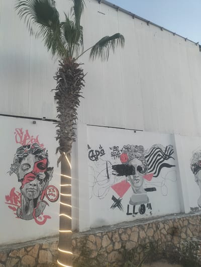 The image captures a section of a white, corrugated building wall adorned with graffiti art. A palm tree, wrapped with festive string lights, stands in front of the wall. The graffiti depicts stylized human faces, one with a red mask-like overlay, and abstract patterns. To the left, a fragmented face is rendered in black, white, and red, with graffiti tags above it. To the right, another stylized face is painted, featuring black scribbles, pink geometric shapes, and a tic-tac-toe game motif. The ground in front of the wall is a stone-laid embankment with sparse vegetation. The setting appears to be outdoors, likely in a public or recreational area. The city of Sidi Abdelrahman, Egypt is mentioned as the location, suggesting a coastal or resort environment. The time of day is likely late afternoon or early evening, given the warm glow of the string lights.
